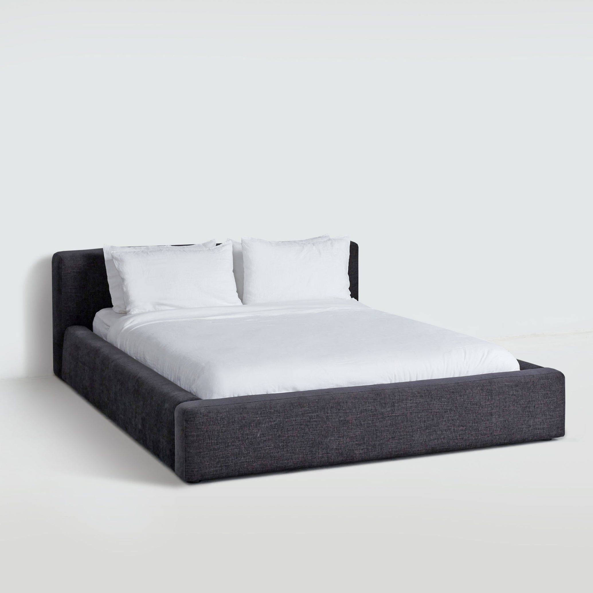 Low-profile upholstered Cosmos Bed in Charcoal, side view, showing wrapped platform frame and cushioned headboard. Low-profile upholstered Cosmos Bed in Charcoal, side view, showing wrapped platform frame and cushioned headboard.