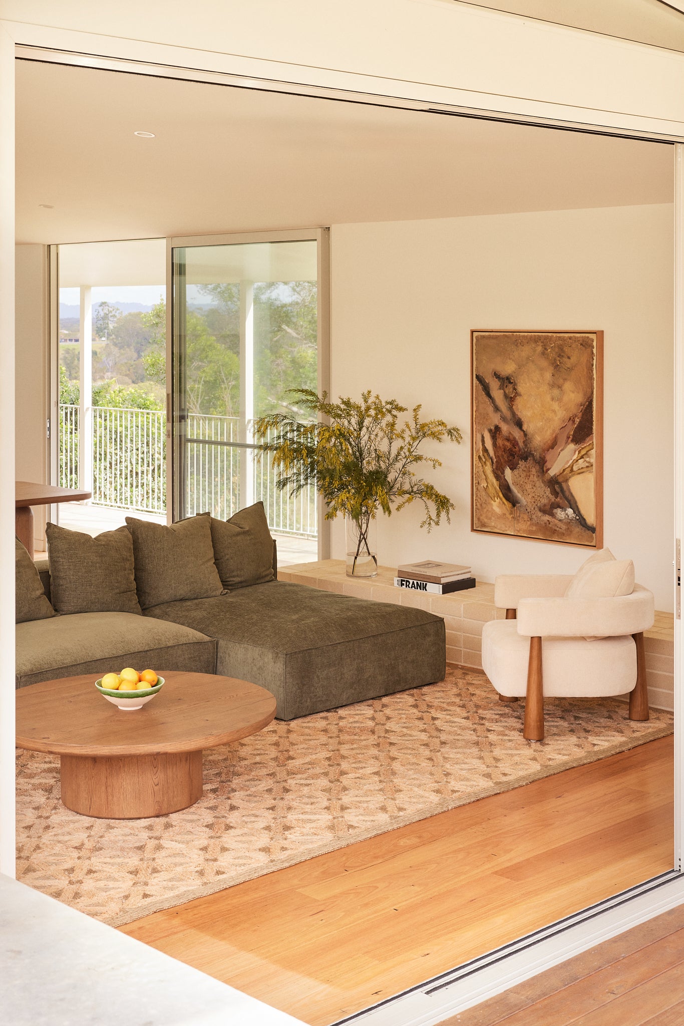 Cosmos sofa with chaise in Forest fabric styled in a contemporary living room with the Alta round coffee table in aged brown Oak, Arlo armchair in oatmeal textured fabric, and original artwork by Stephen Skinner.